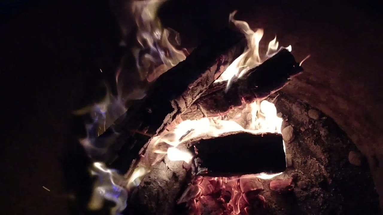 Bright flames curl around stacked dark wood above glowing embers.