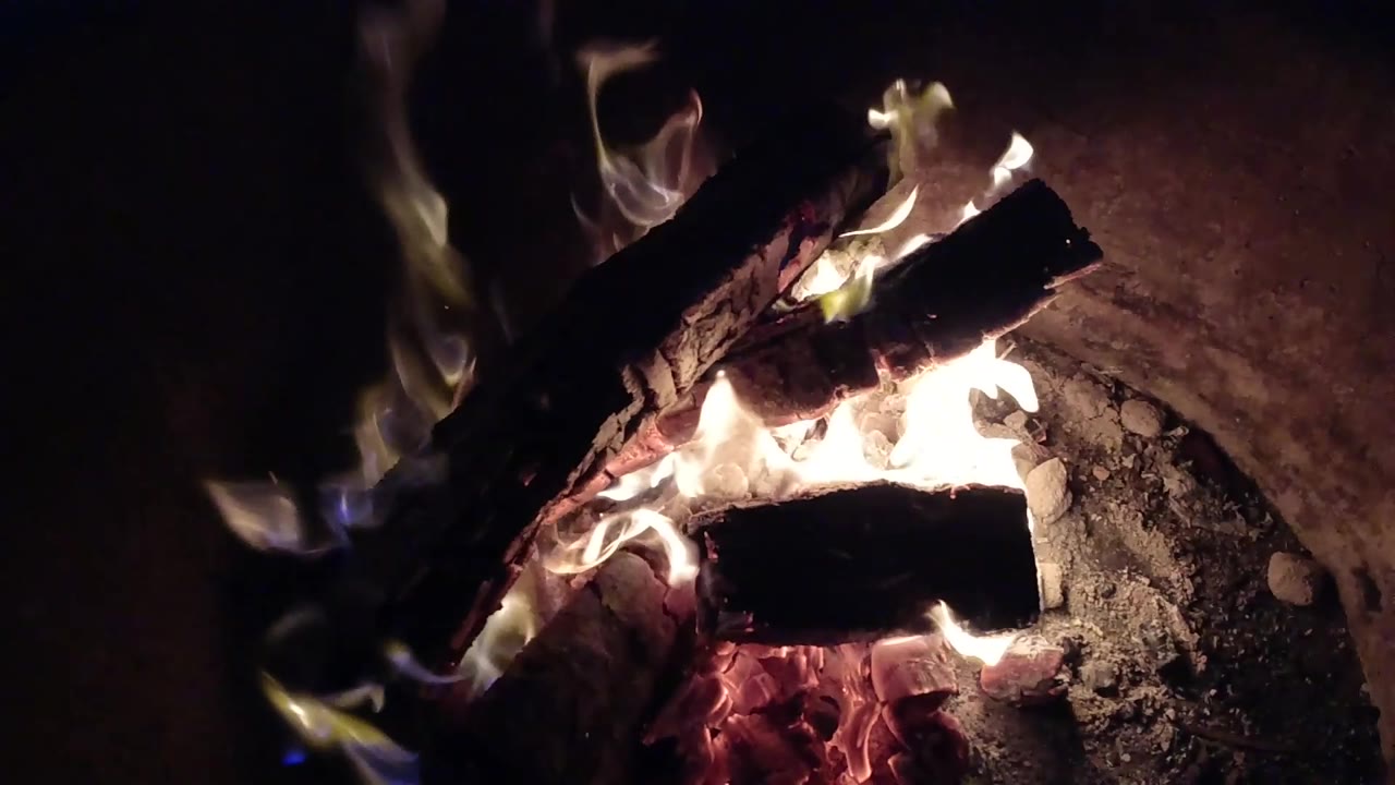 At 0:02, angled burning logs sit over a bright bed of embers in Pismo Beach, California, with flame tips leaning upward and no visible people.