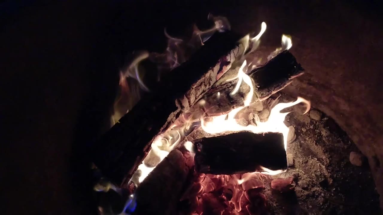 Bright flames curl around the lower and upper logs in the Pismo Beach fire pit, with red embers concentrated near the bottom and no visible people.