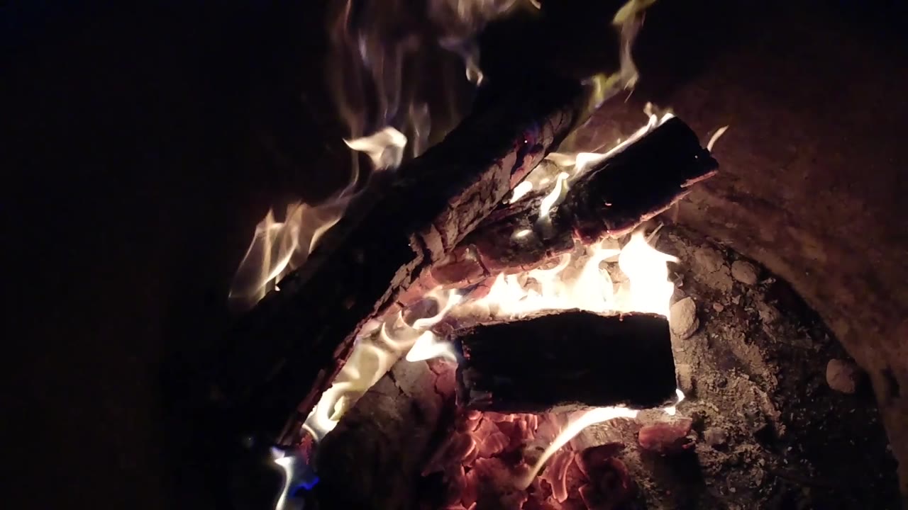 A bright flame crosses the front log in the Pismo Beach fire pit while the upper log stays dark and charred, with no visible people.