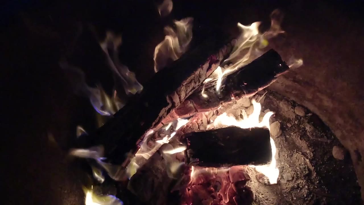 At 0:00 in Pismo Beach, California, charred logs burn over red embers with yellow-white flames rising from the lower edge and no visible people.