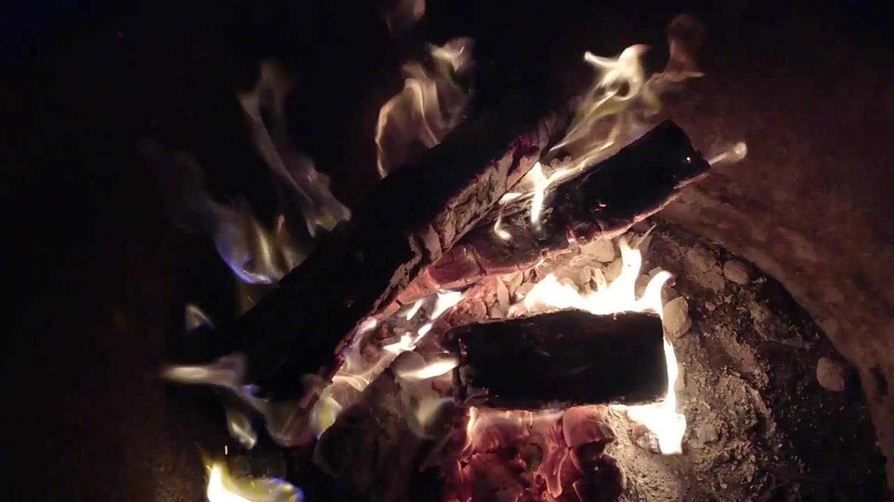 At 0:01 in Pismo Beach, California, pale flames curl around two blackened logs while red coals glow below and the surrounding area stays dark, with no visible people.
