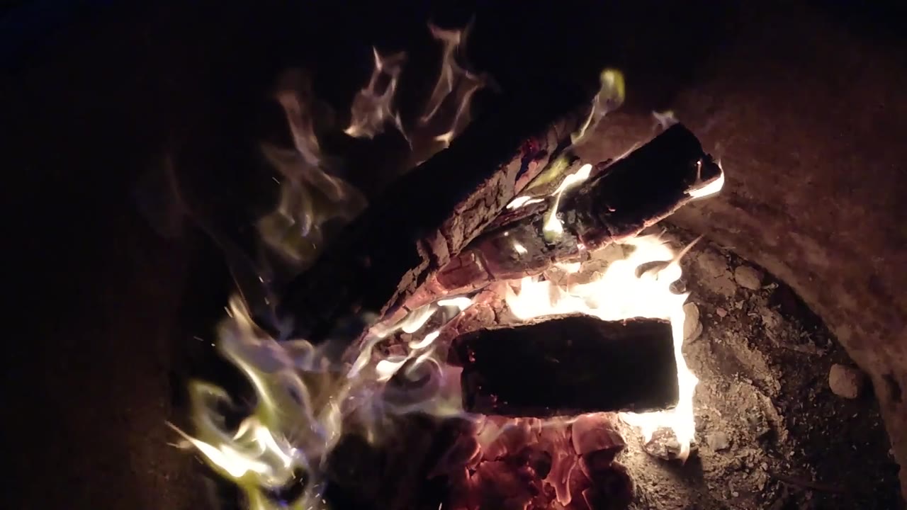 At 0:02 in Pismo Beach, California, bright flames flare along the right side of crossed charred wood, with red embers visible underneath and no visible people.
