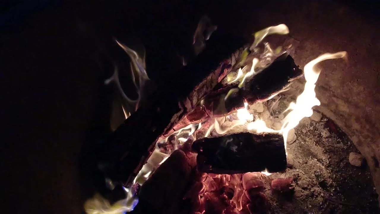 Pismo Beach, California fire close-up with a bright flame curling above charred wood, ember glow below, and no visible people.