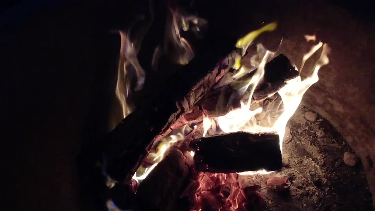 Pismo Beach, California close fire view with crossed logs, bright flames wrapping the wood, red embers underneath, and no visible people.