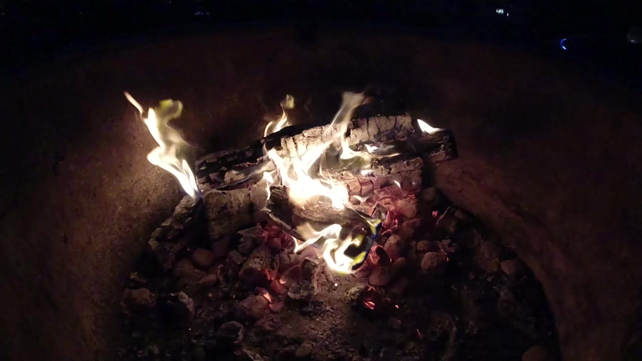 At 0:00, bright flames burn across charred logs and red embers in a dark Pismo Beach campfire scene, with no visible people.