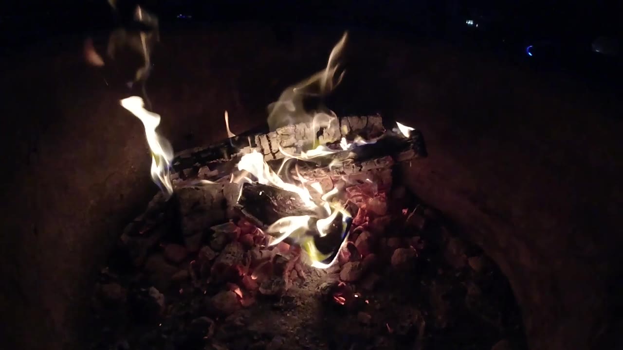 At 0:02, taller flames lift from the ember bed around blackened wood inside the dark Pismo Beach fire pit, with no visible people.
