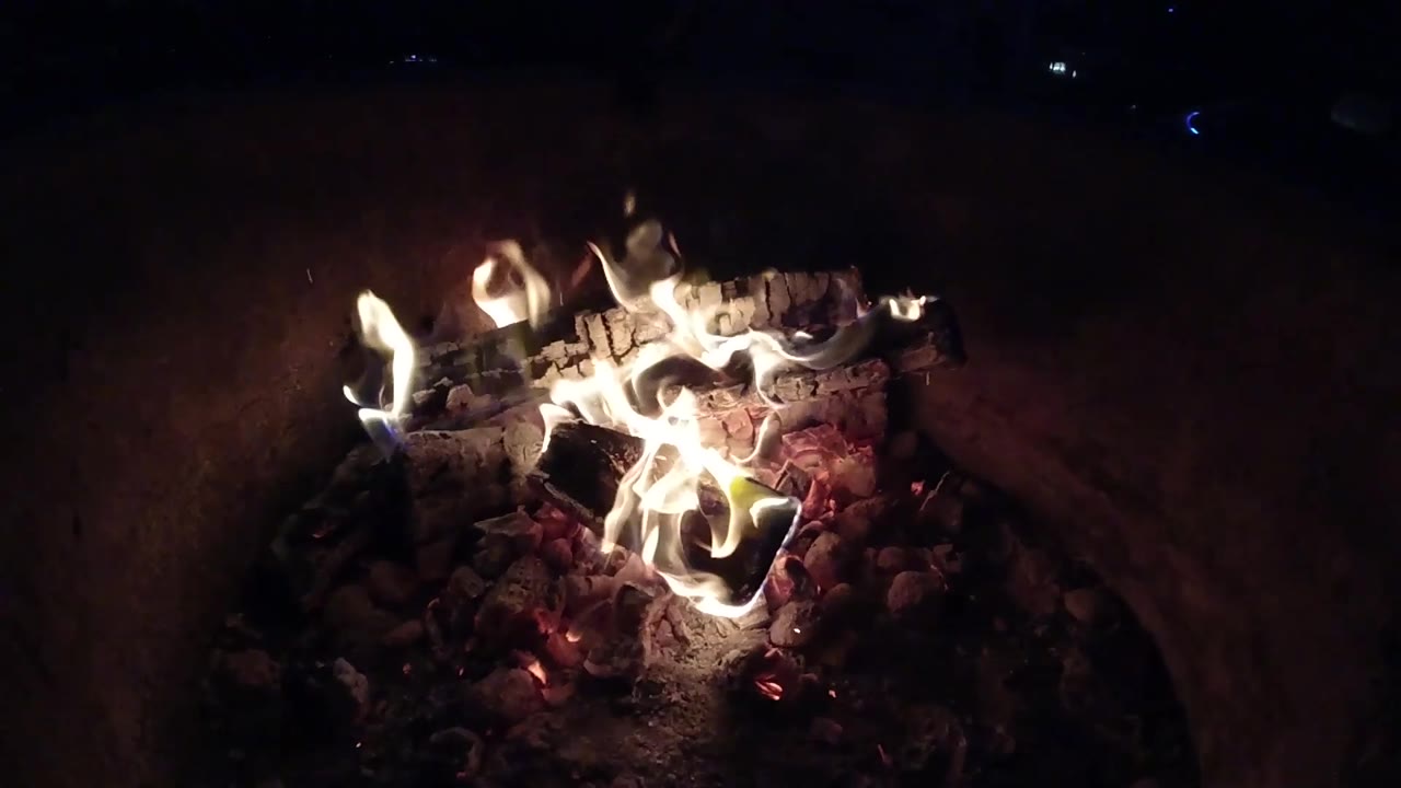 At 0:03, bright low flames spread across the coals and charred wood in the Pismo Beach campfire, with the surrounding area mostly black and no visible people.
