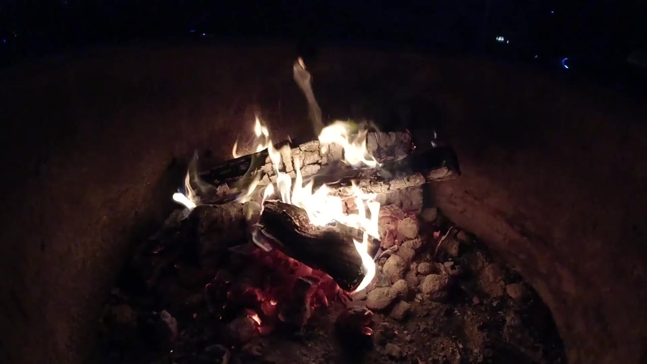 At 0:01, pale flames rise higher over the charred logs and ash in the Pismo Beach fire container, with no visible people.