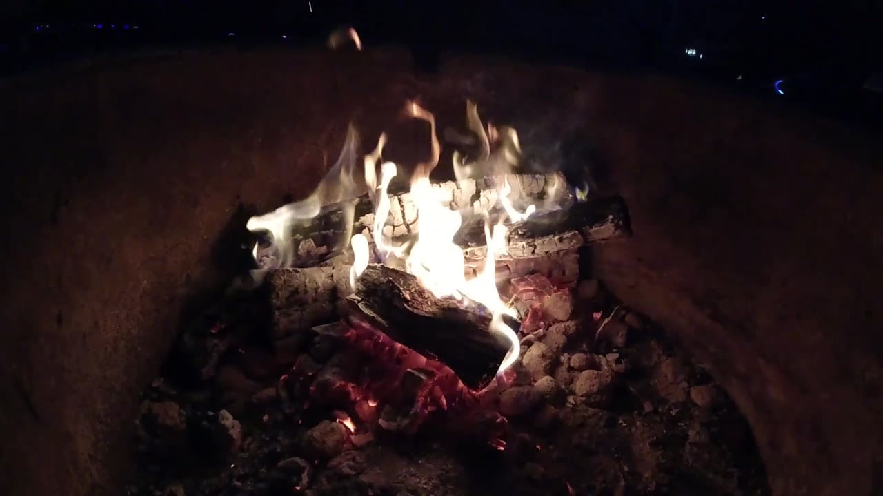 At 0:02, the fire burns brighter across the stacked logs in the Pismo Beach container, with red embers visible below and no visible people.