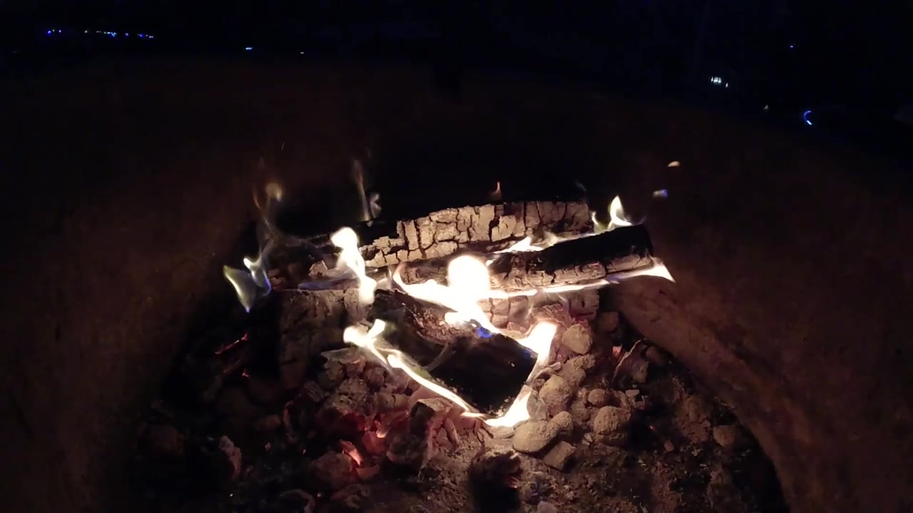 At 0:02, the campfire remains centered with a long upper log, glowing coals, and a dark sandy background in Pismo Beach, with no visible people.