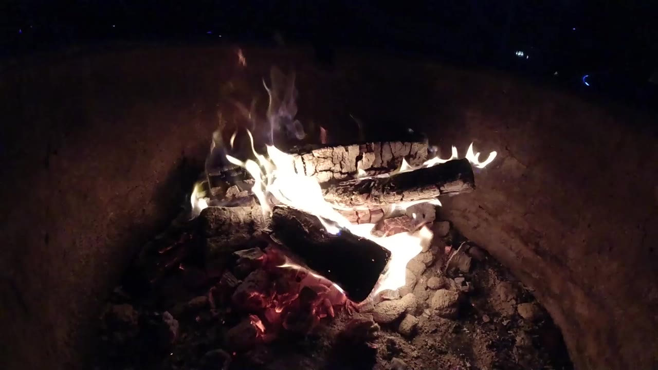 A low burning campfire in Pismo Beach, California shows split logs over ash and red embers, with flames rising on the left side and no visible people.