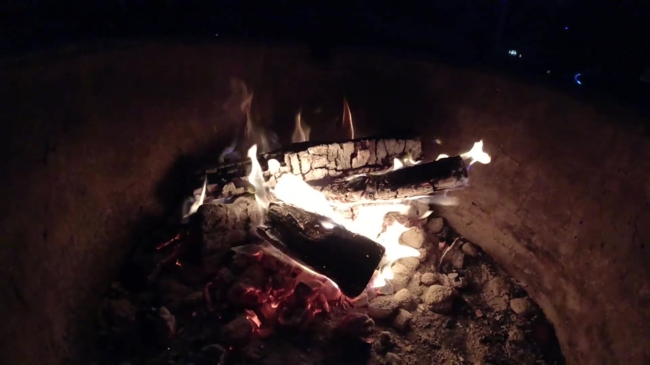 A nighttime campfire in Pismo Beach, California shows curled flames around charred logs, scattered ash, red coals below, and no visible people.