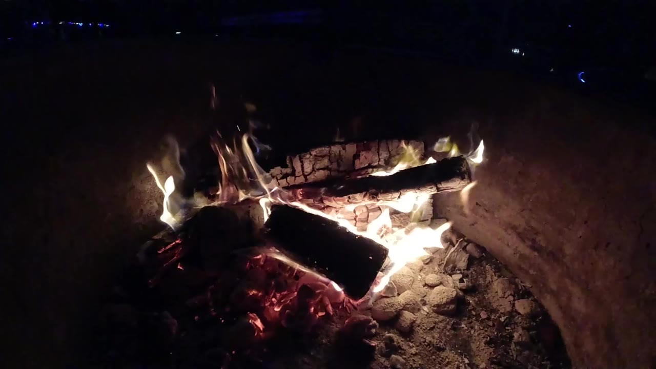At 0:01 in Pismo Beach, California, flames curl around stacked logs while red embers glow beneath them and no people are visible.