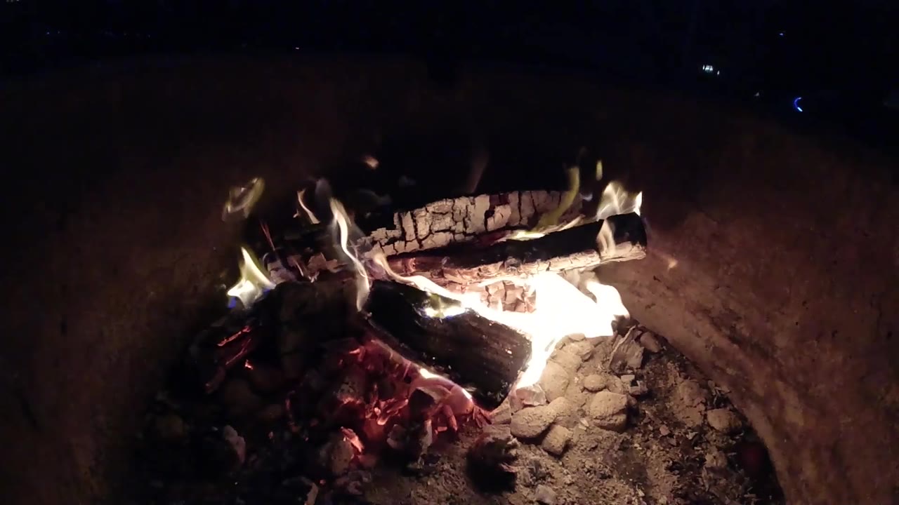 At 0:02 in Pismo Beach, California, bright firelight washes over charred logs in the pit while the surrounding area stays mostly black, with no visible people.