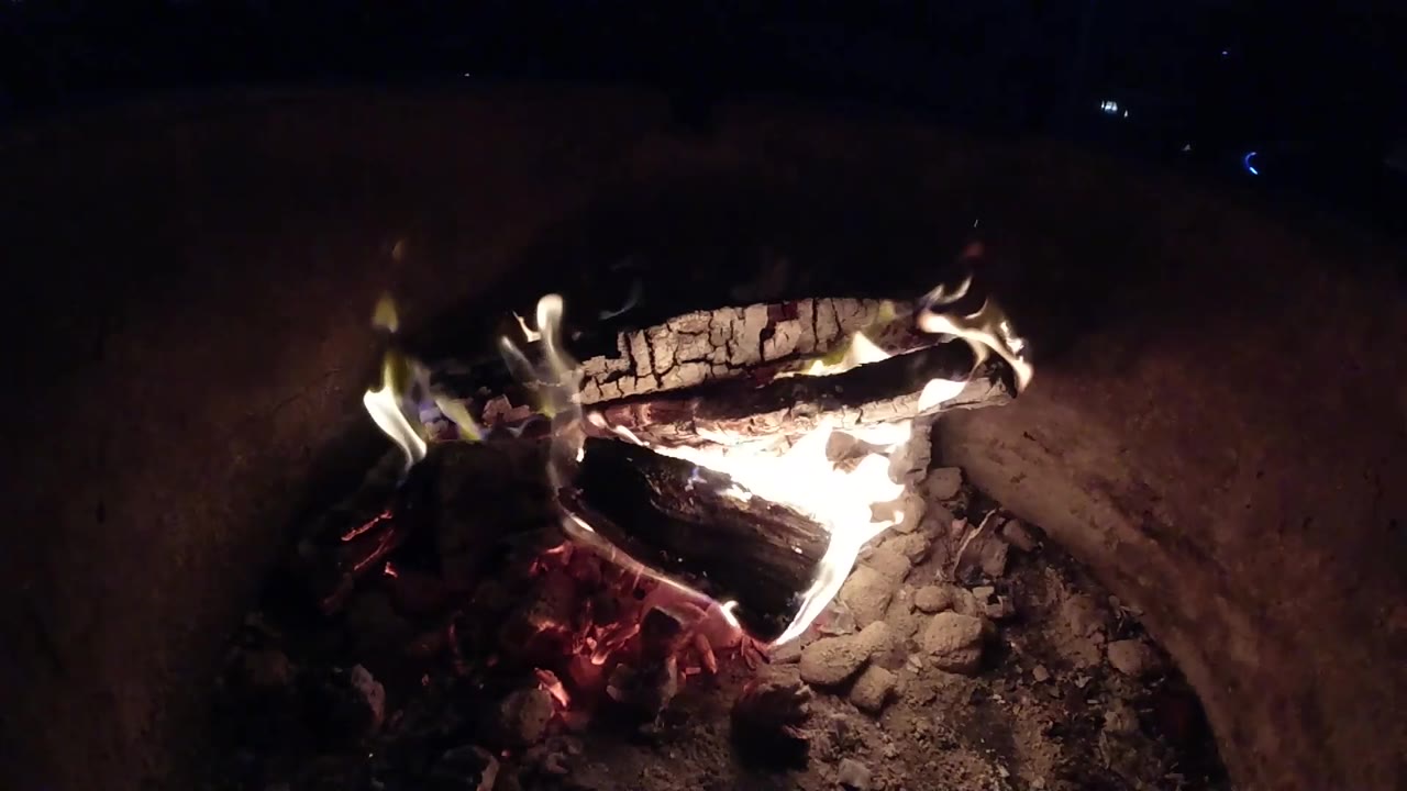 At 0:03 in Pismo Beach, California, the fire shifts lower in the frame with glowing coals on the left and burning logs across the center, with no visible people.