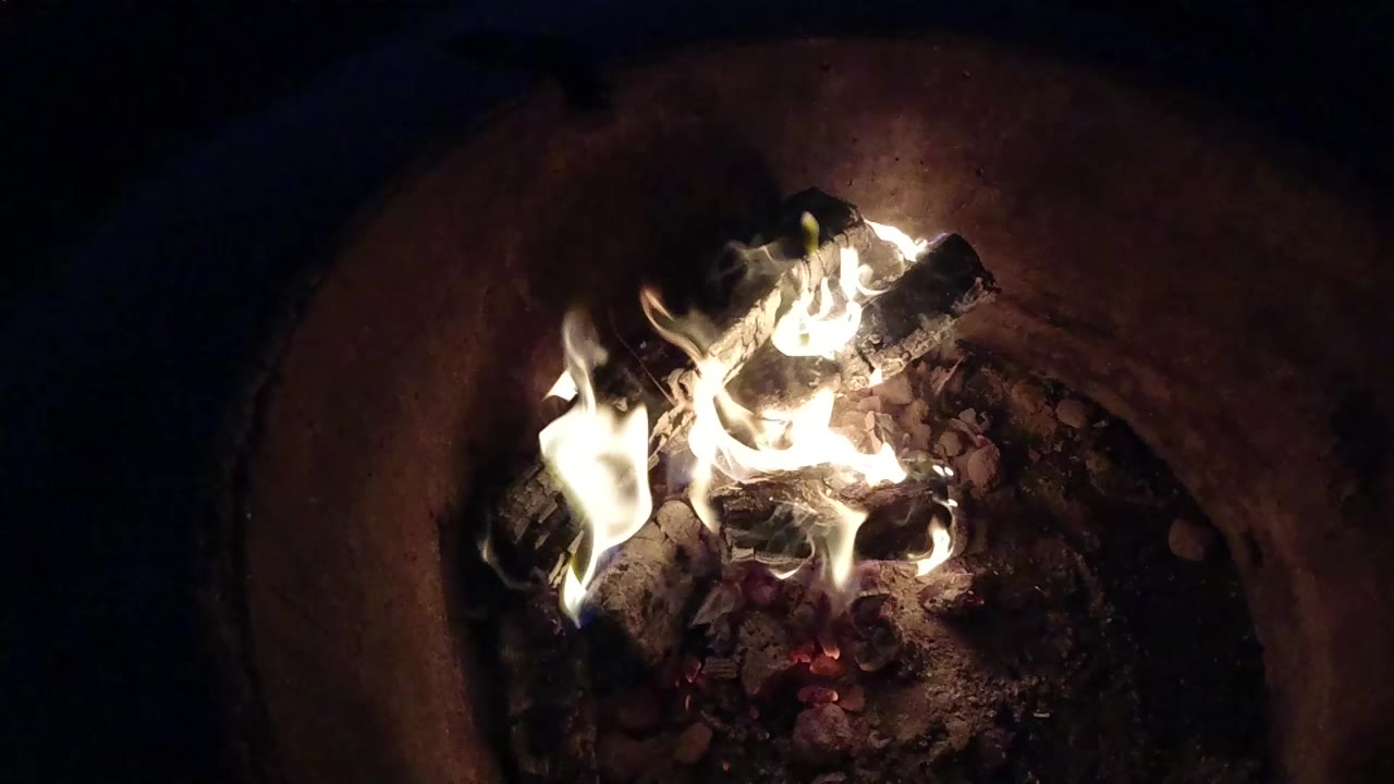 At 0:00, flames rise from charred logs and red embers in a dark round fire pit in Pismo Beach, California, with no visible people.