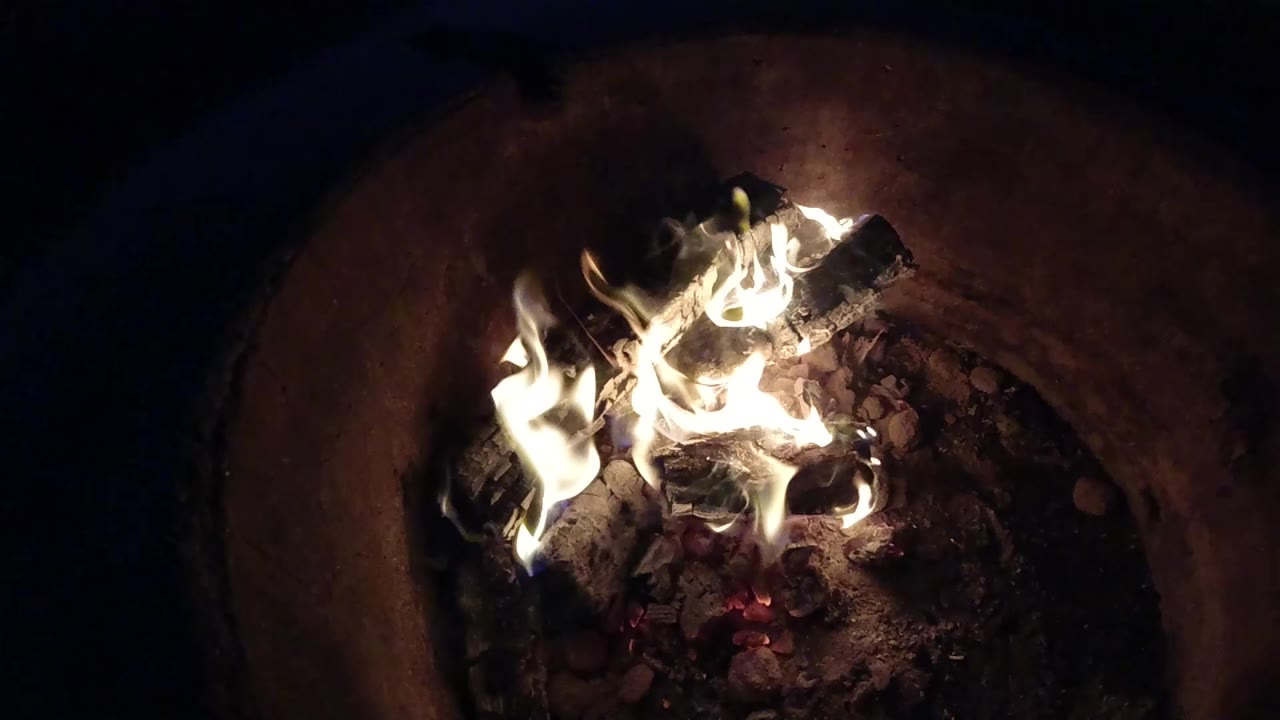 At 0:01, the fire burns brighter across the center logs, with pale flames, ash, and embers visible in Pismo Beach, California, and no visible people.
