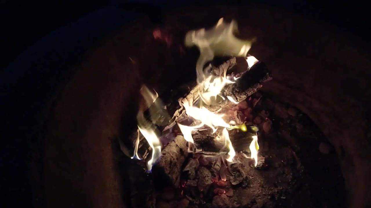 At 0:03, taller flames lift above the charred logs and expose glowing red coals underneath in the Pismo Beach, California backyard fire, with no visible people.