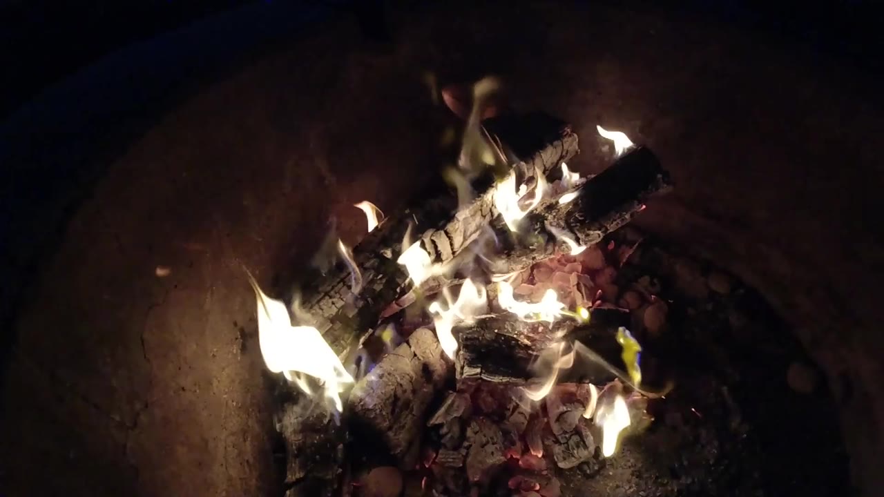 At 0:01, flames brighten along the left and upper logs while red embers sit beneath them in the Pismo Beach fire pit, with no visible people.