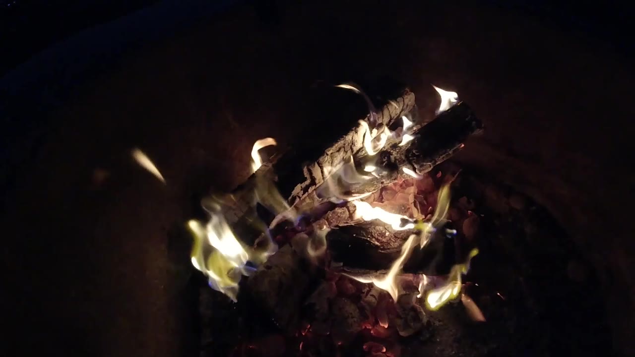At 0:02, the burning logs sit farther right in the frame with smaller flames and deep shadow around the rounded fire pit, with no visible people.