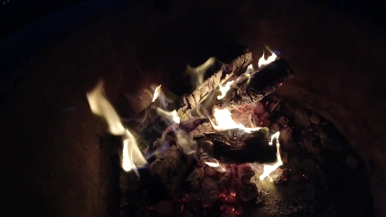 At 0:03, taller yellow flames flare in the foreground in front of charred logs and glowing embers in Pismo Beach, California, with no visible people.