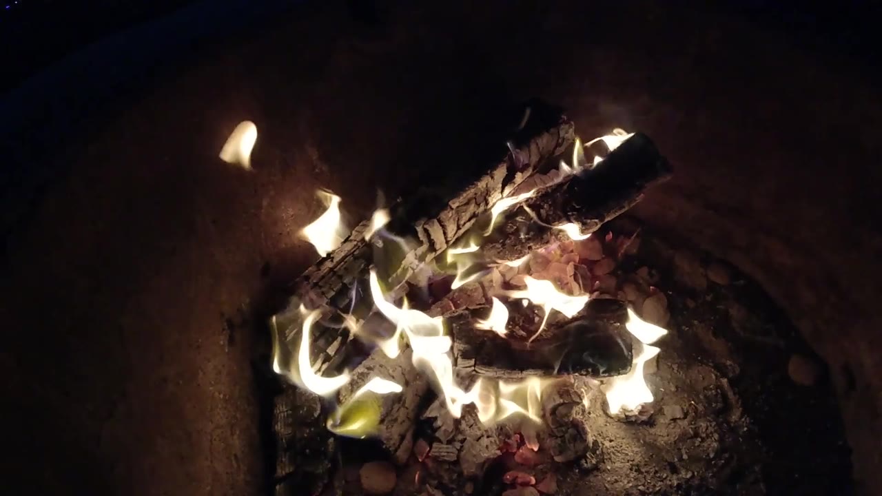 At 0:02, the fire spreads wider across the ash bed while a dark charred log sits near the top of the Pismo Beach fire pit, with no visible people.