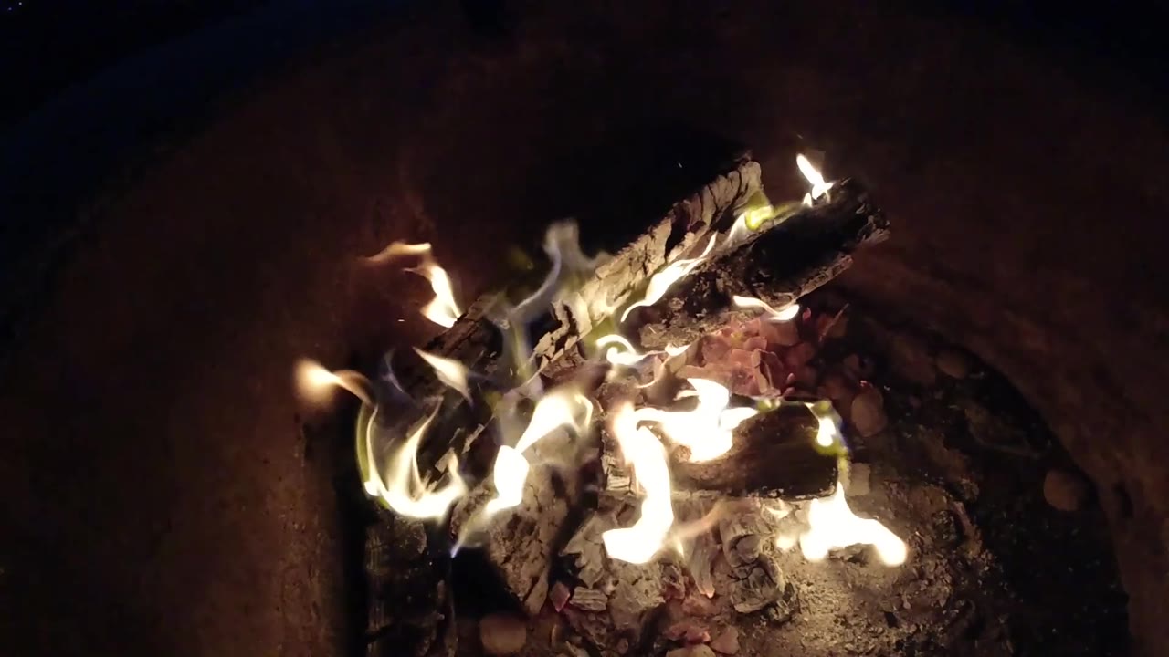 At 0:03, bright white-yellow flames flare beneath blackened logs and red embers in the Pismo Beach campfire, with no visible people.