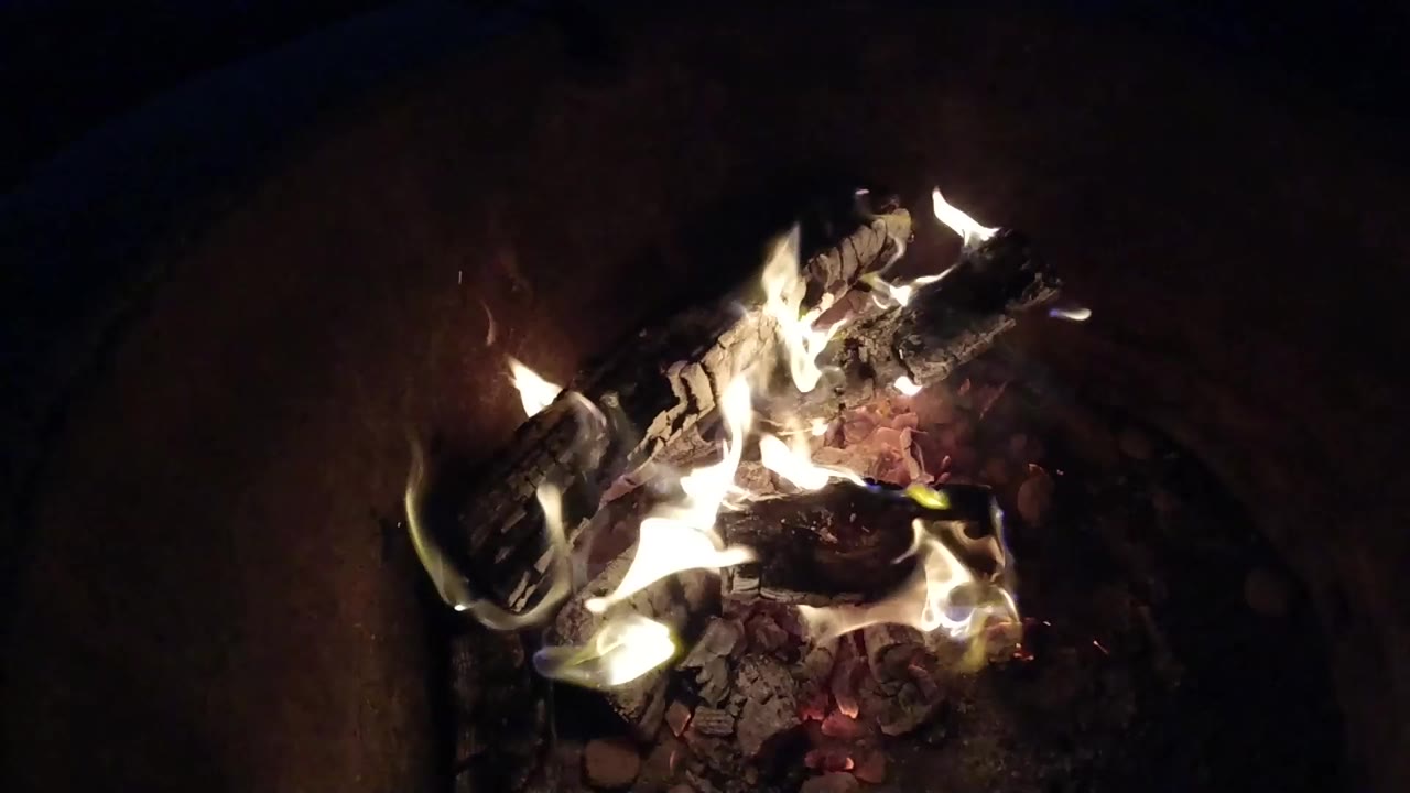 Close-up of pale flames curling around charred wood, ash, and red embers in Pismo Beach, California, with no visible people.