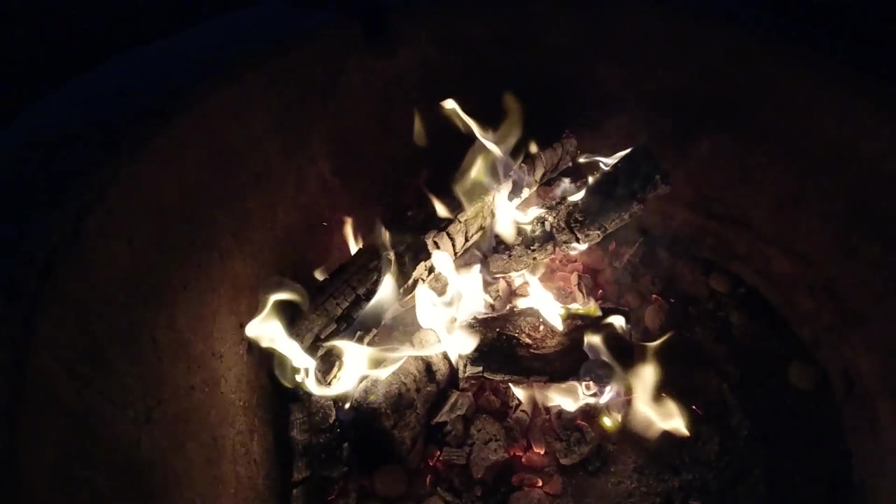 A brighter flare spreads across the top of the burning logs and ash inside a dark fire pit in Pismo Beach, California, with no visible people.