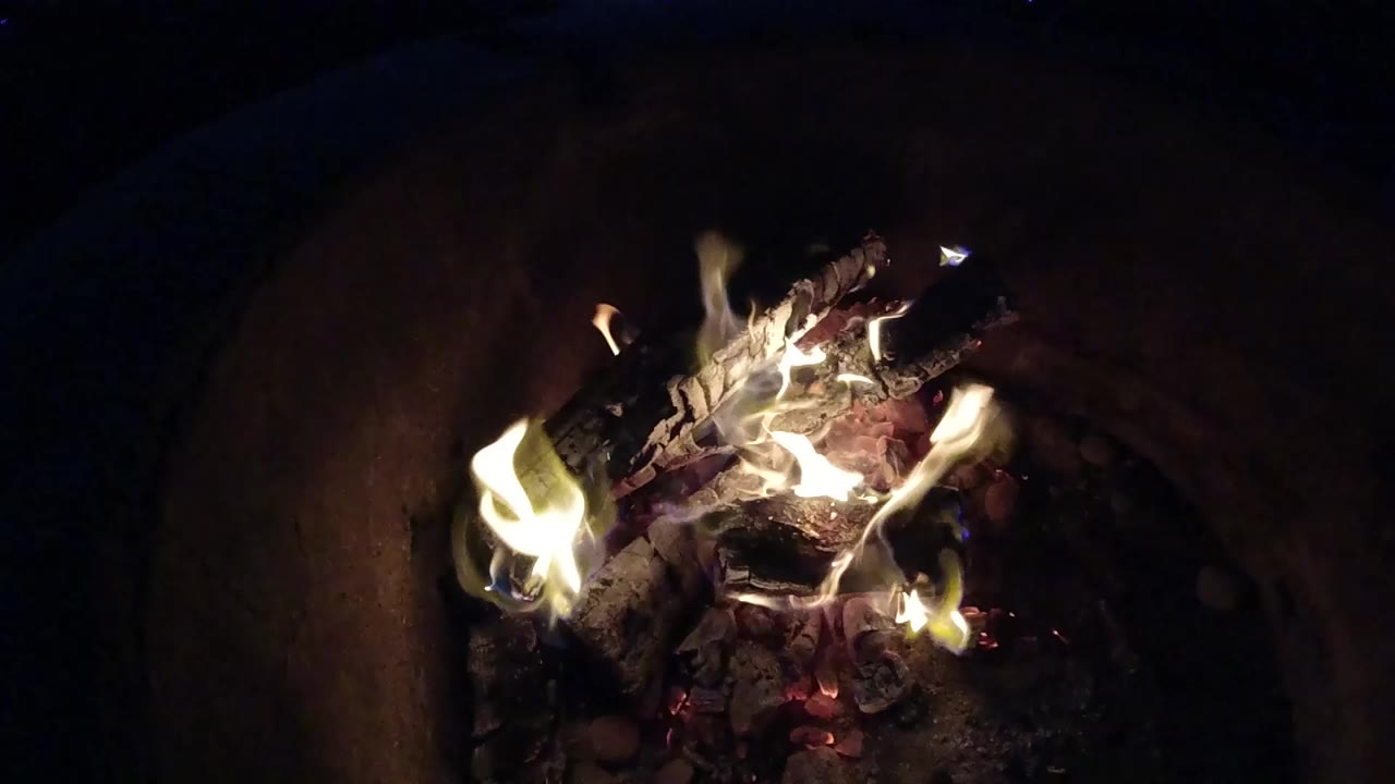 At 0:01, white yellow flames curl higher over charred logs in the deep container, with glowing coals below and no visible people.