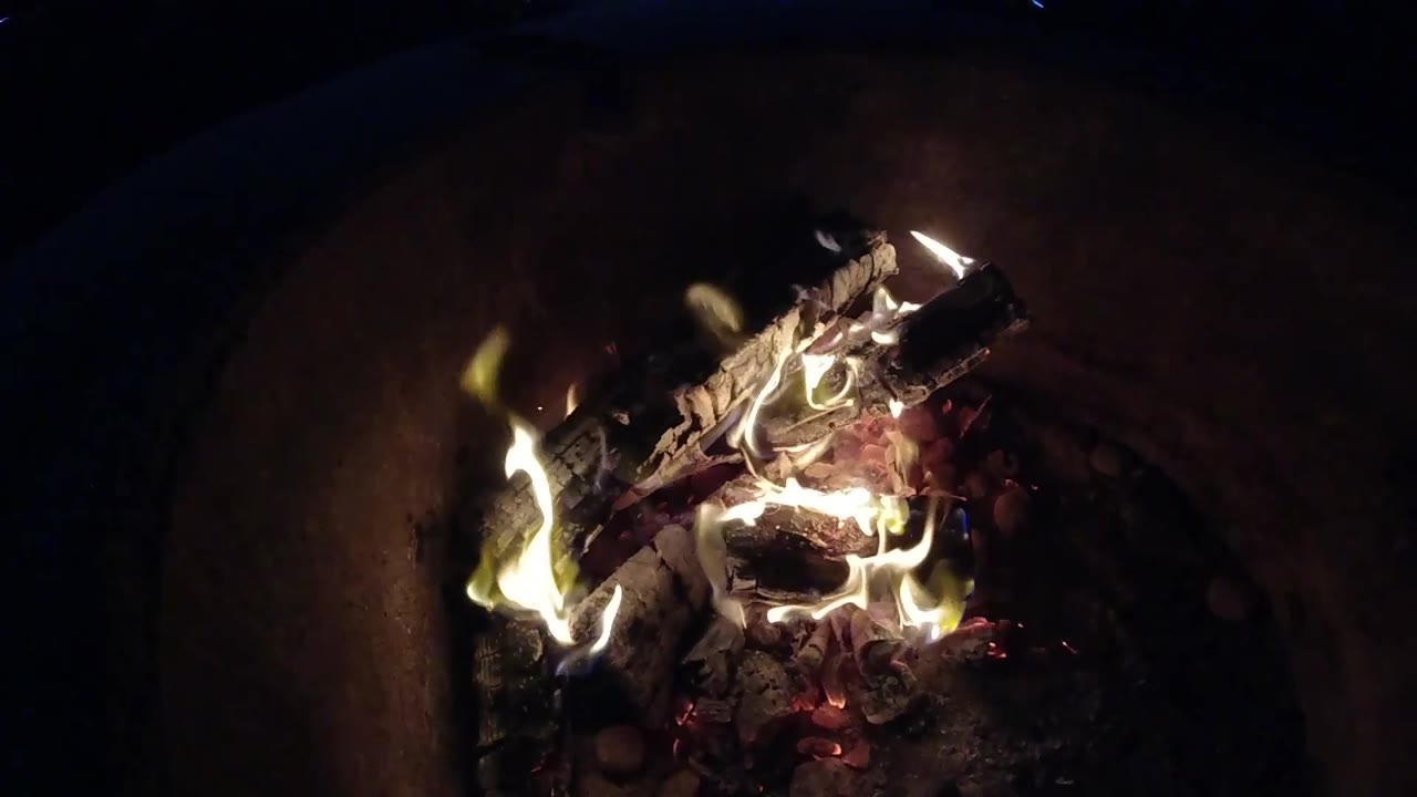 At 0:02, the main flame shifts toward the left side of the logs while embers glow beneath the wood in the Pismo Beach clip, with no visible people.