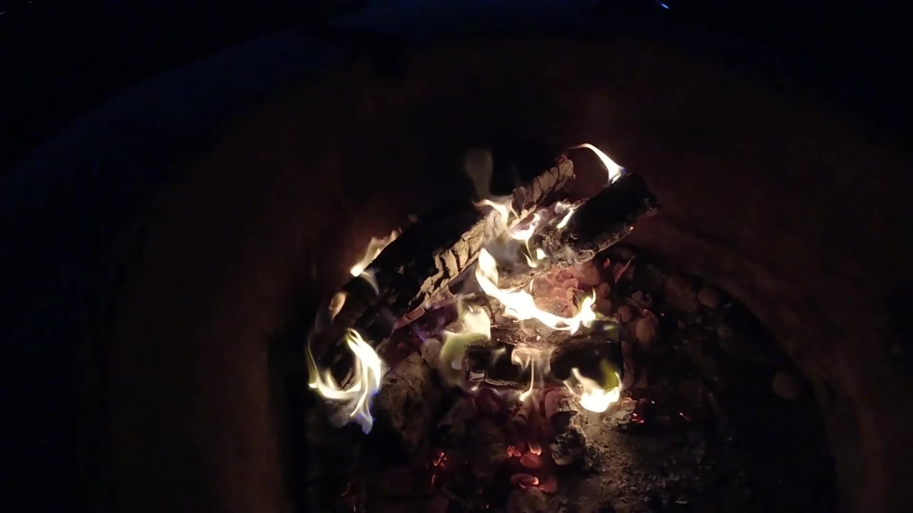 At 0:03, lower flames wrap around the front and center of the firewood, with a dark rounded container wall behind and no visible people.