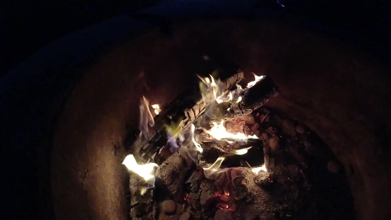 At 0:00, burning logs glow inside a round fire pit in Pismo Beach, California, with bright flames rising from the left side and no visible people.