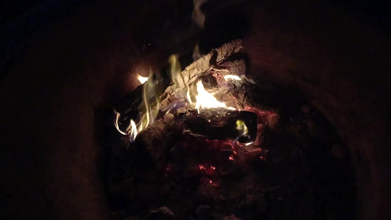 At 0:00, burning logs glow inside a dark recessed fire pit in Pismo Beach, California, with small flames rising and no visible people.