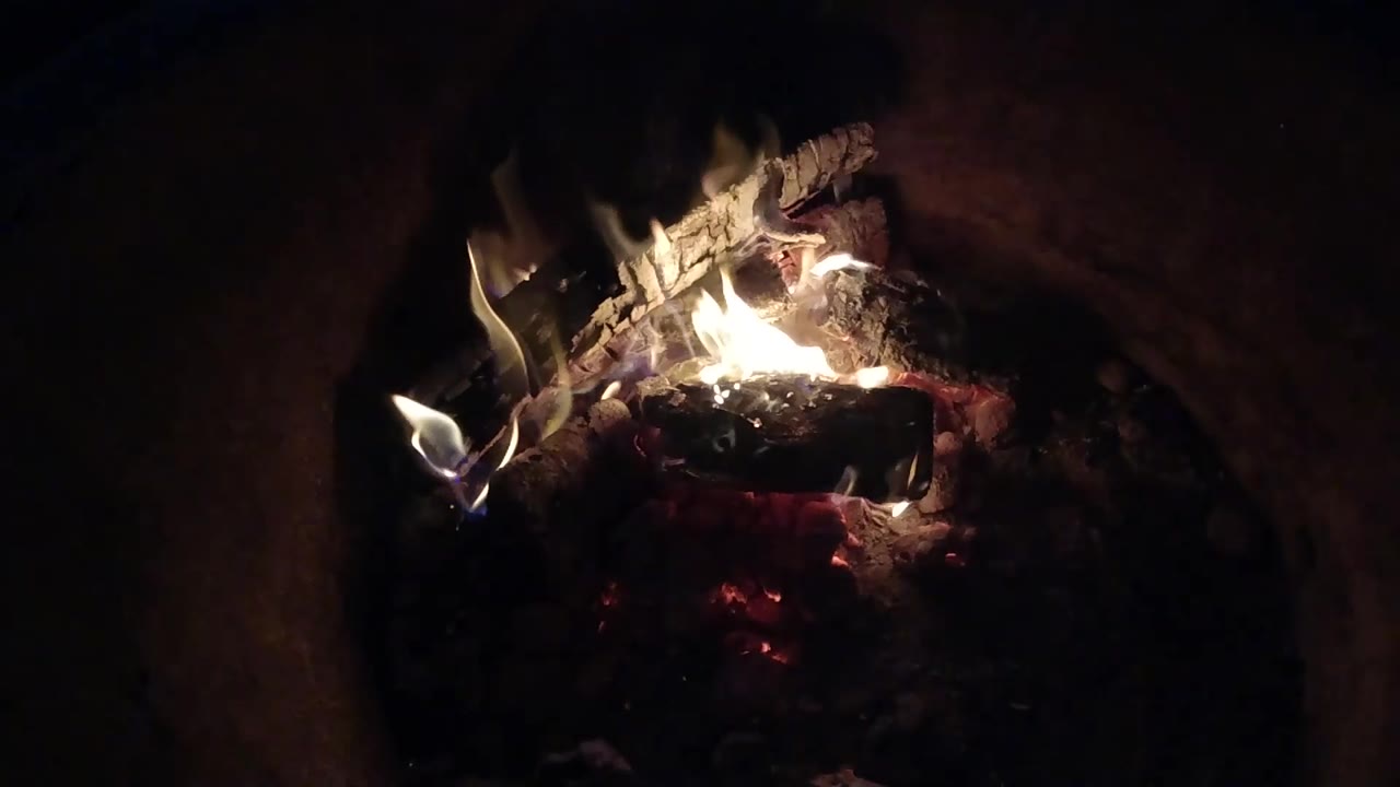 At 0:02, the fire sits lower across the wood, with a bright flame on the left edge and glowing coals underneath, no visible people in frame.