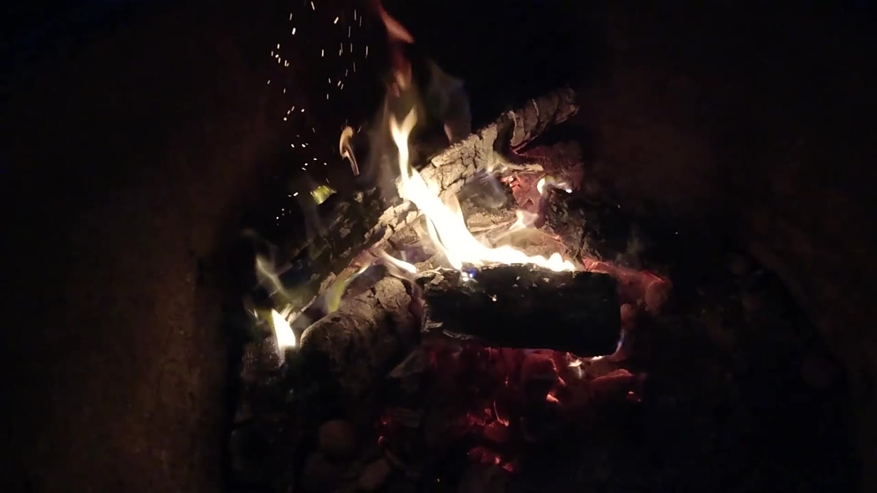 Small sparks rise above bright flames and charred logs in the dark Pismo Beach fire pit, with no visible people in this frame.