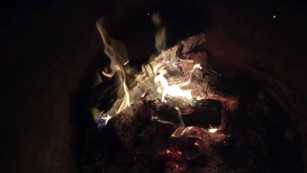 Bright white-yellow flames curl across charred wood and glowing coals in Pismo Beach, California, with deep shadow around the fire and no visible people.