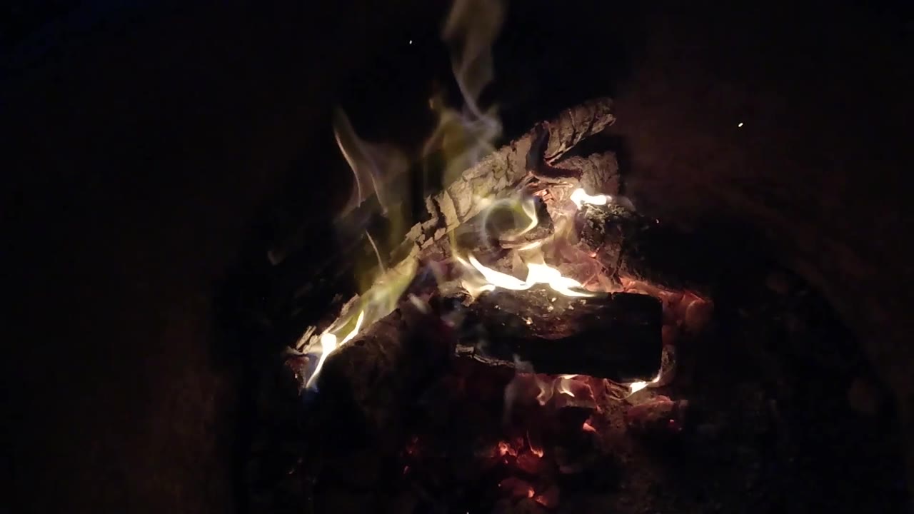 Thin smoke and pale flames rise from blackened logs and ember pockets in Pismo Beach, California, with open darkness to the right and no visible people.