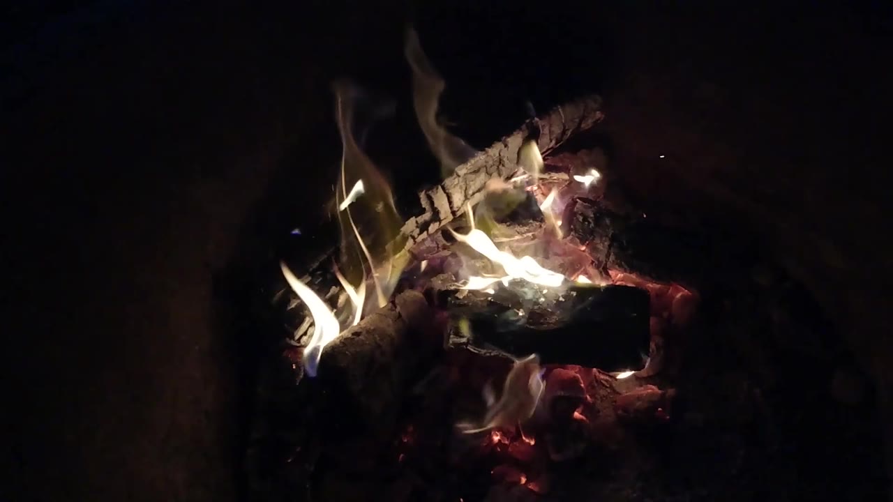 Flames lean across a compact bed of embers and charred wood in Pismo Beach, California, with the fire positioned left of center and no visible people.