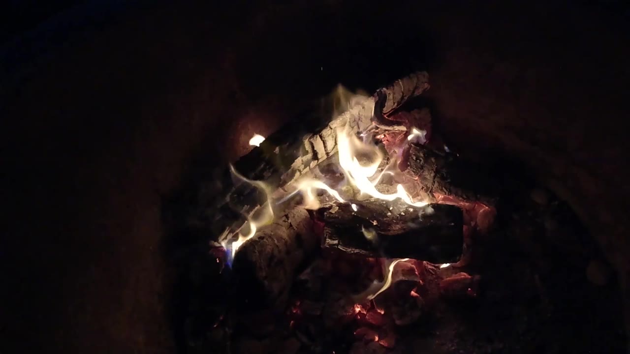 At 0:03, low flames spread across the center of blackened wood and red embers in the Pismo Beach, California clip, with no visible people.