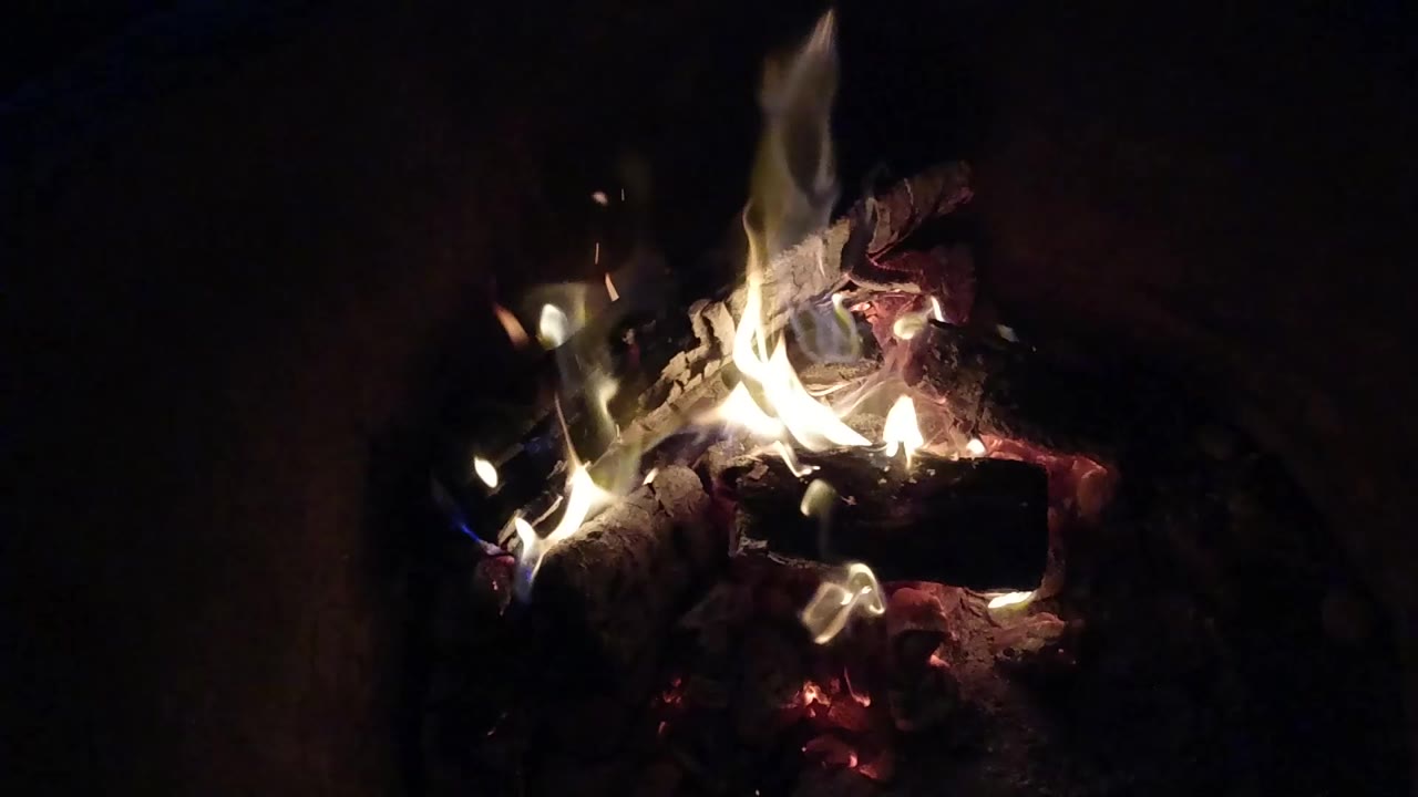 At 0:00, small flames rise over charred logs and red embers in a dark fire pit in Pismo Beach, California, with no visible people.