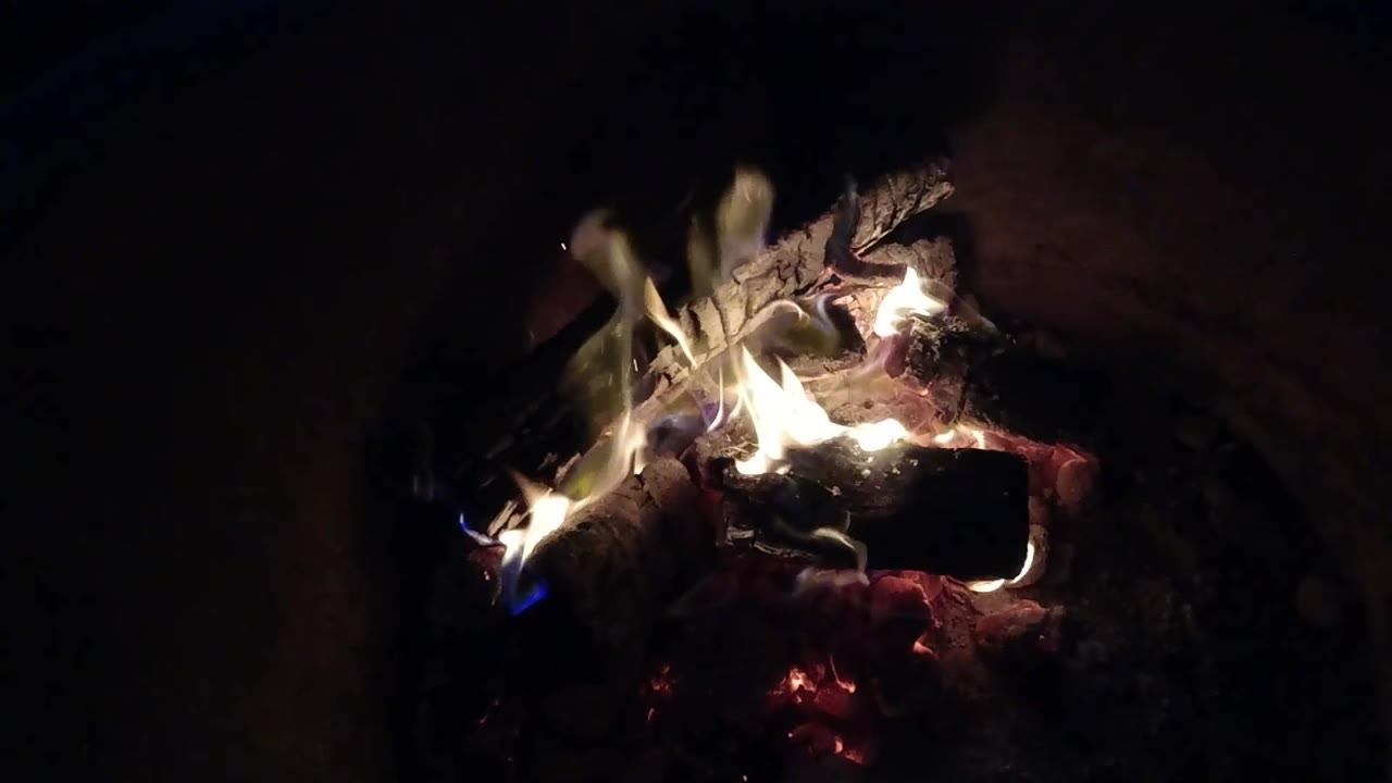 At 0:02, bright firelight gathers on the right side of the charred logs in Pismo Beach, California, with embers visible below and no visible people.