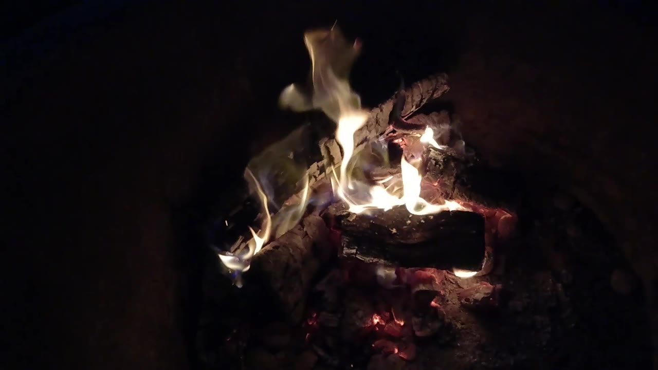 At 0:03, pale flames curl along the left side of a blackened log in the Pismo Beach fire pit, with red embers underneath and no visible people.