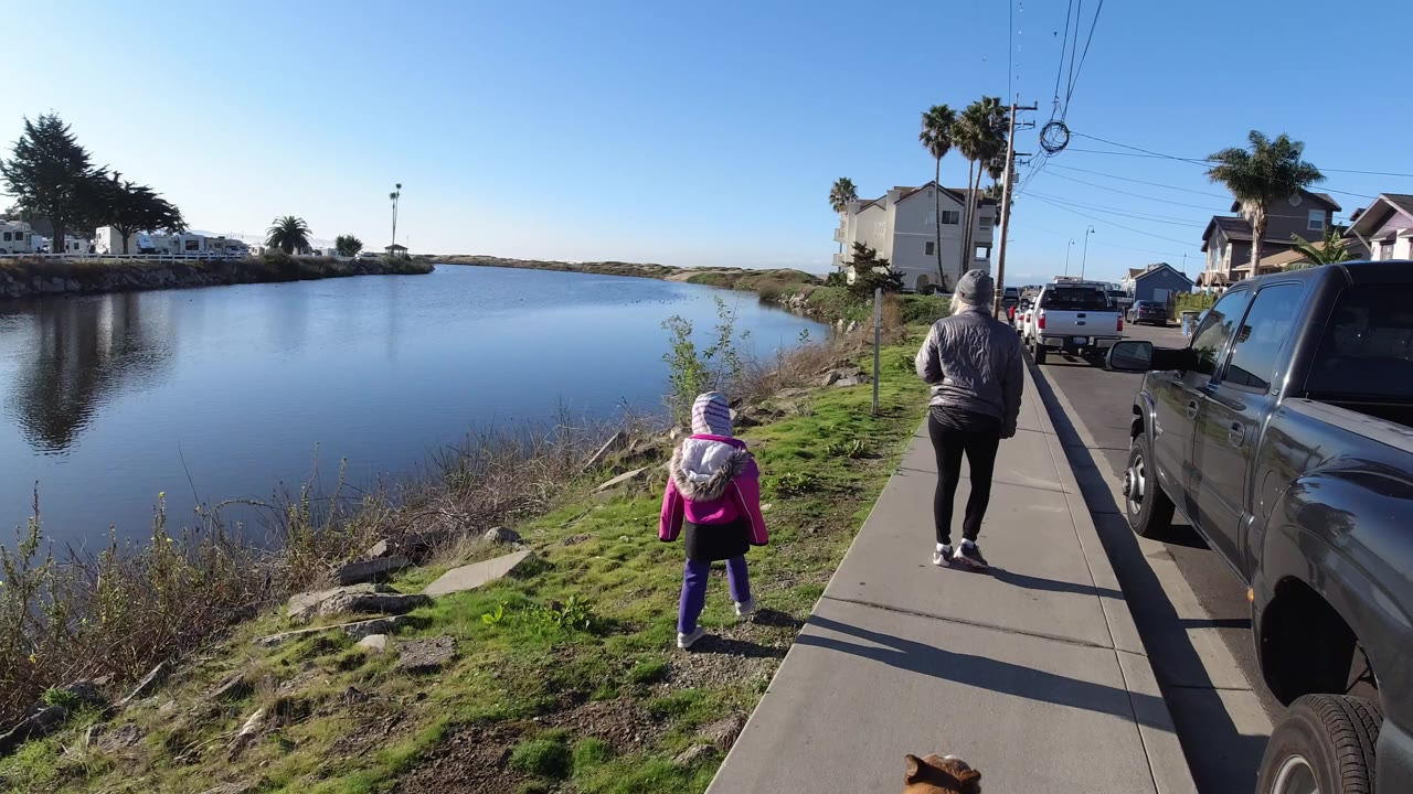 In Pismo Beach, a child in a pink jacket and striped hat walks behind an adult on a sidewalk beside still water, with parked vehicles and homes along the street.