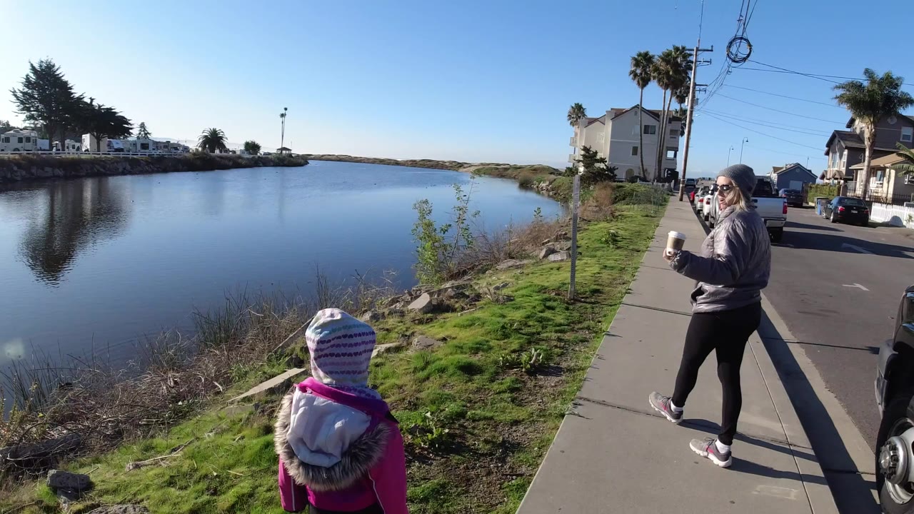At Pismo Beach, a woman holding a cup walks on a sidewalk while a child in a striped hat faces the blue water beside grass and shoreline rocks.