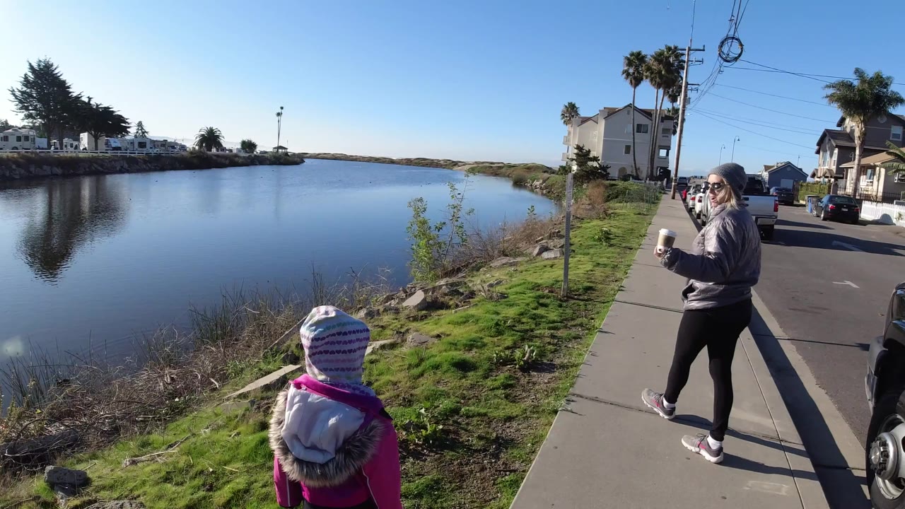 At Pismo Beach, the child remains in the foreground near the grassy water edge while the woman with a cup is farther along the sidewalk beside parked vehicles and homes.