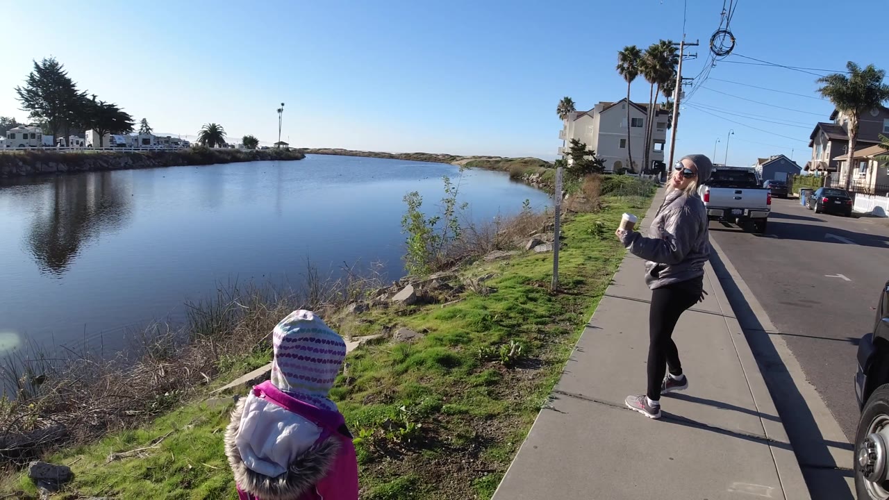 At Pismo Beach, the woman in sunglasses and gloves stands on the sidewalk holding a cup while the child faces the water, with bright sun lighting parked cars and houses behind them.