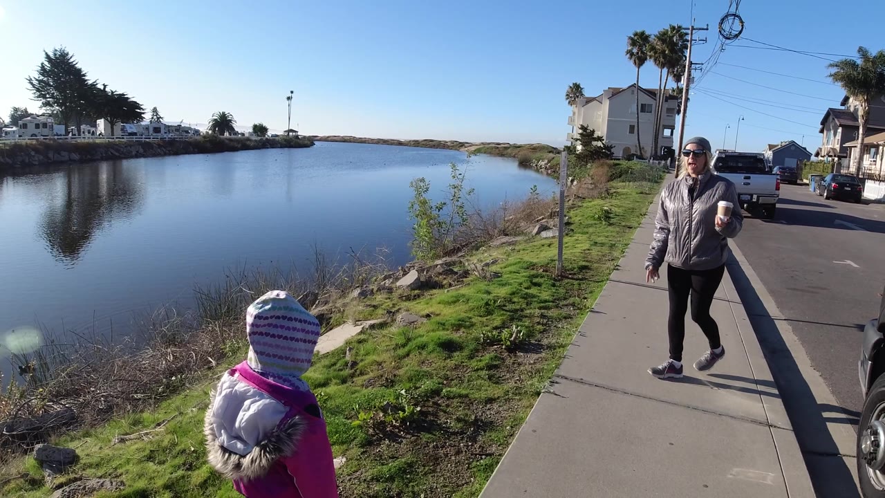 Wide view of blue water with a child in the foreground and woman on the sidewalk.