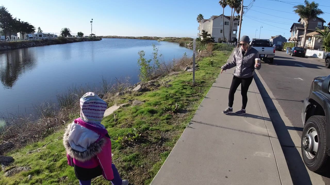 In Pismo Beach, California, a woman holding a cup stands ahead on a sidewalk while a child in a pink jacket walks near the grassy bank beside still water.